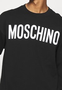 Sweat-shirt noir avec le texte blanc audacieux "MOSCHINO" sur la poitrine. Design classique à col rond avec poignets et ourlet côtelés. Texture douce.