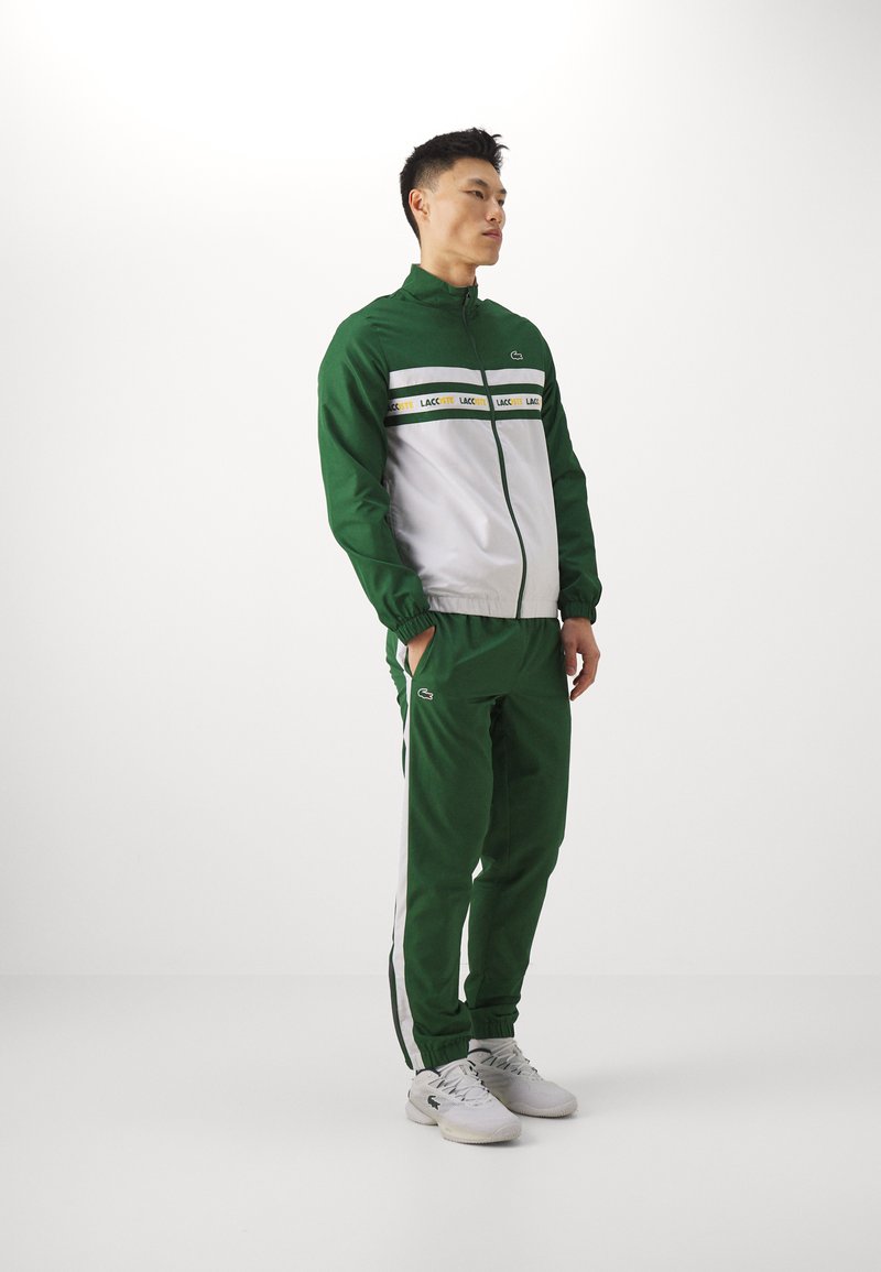 Survêtement vert avec une veste blanche à zip, ornée de rayures horizontales et d'accents jaunes. Tissu léger avec des poignets et une taille élastiques.