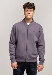 Fioletowa bluza z zamkiem błyskawicznym i stójką, z prążkowanymi mankietami i dołem, z małym logo na klatce piersiowej. Gładka, miękka tekstura materiału.