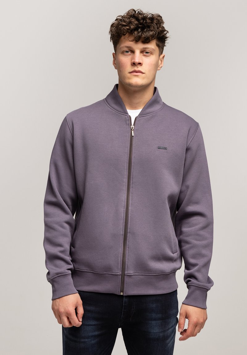 Fioletowa bluza z zamkiem błyskawicznym i stójką, z prążkowanymi mankietami i dołem, z małym logo na klatce piersiowej. Gładka, miękka tekstura materiału.