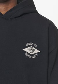 Sudadera azul marina con un logo en forma de diamante blanco en el pecho que dice "PEGADOR No. 1" y "Construido para durar Pgr".