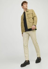 Beige Jacke mit mehreren Taschen, schwarzes Hemd darunter, hellfarbige Hose und schwarze Slipper. Glatte Textur; lässiger Stil.