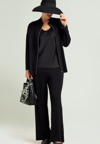 Zwarte gebreide cardigan over een zwart top, gecombineerd met zwarte flare pants. Het model houdt een zwarte tas vast met een groene stippen sjaal als accent.