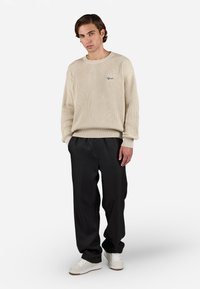 Young man wearing a beige ribbed sweater, black loose pants, and white sneakers standing with hands in pockets against a plain background.