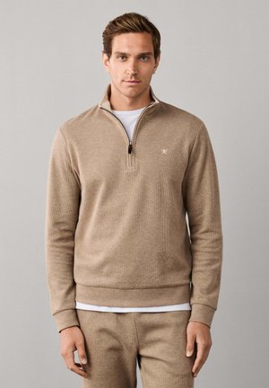 Man wearing beige quarter-zip pullover and matching pants standing against a plain light gray background.
