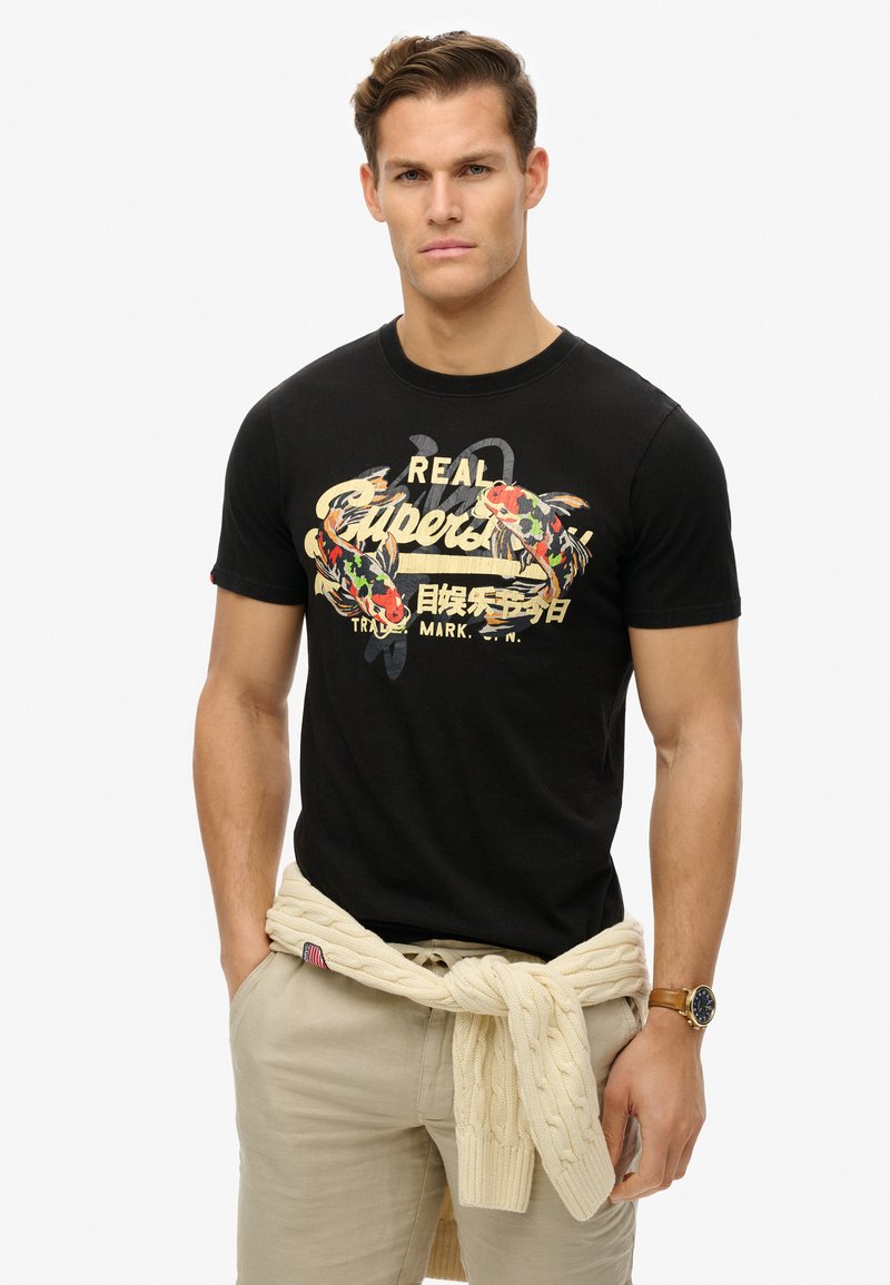 Camiseta de algodón negra con un gráfico de pez colorido y texto. Combinada con pantalones beige claros y un suéter de punto crema anudado a la cintura.