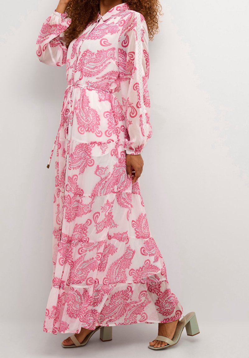 Femme portant une longue robe blanche avec des motifs cachemire roses et des manches longues, associée à des sandales à talons blocs vert clair.