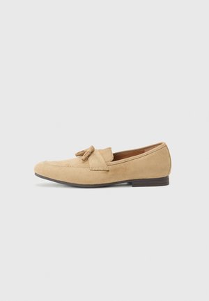 Beige suede loafers with a rounded toe, tassel accent, and stitched detailing. Low, flat brown sole. Simple design without patterns.