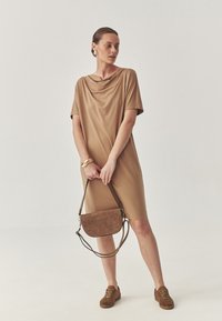 Tan-värinen draped-mekko, jossa on lyhyet hihat ja sileä rakenne. Ruskea mokkanahkainen olkalaukku, jossa on kaareva muoto ja säädettävä kantohihna. Ruskeat lenkkarit.