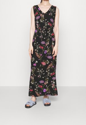 Mouwloze zwarte maxi-jurk met kleurrijk bloemenpatroon en strik aan de voorkant, gedragen met lichtblauwe sandalen met open tenen op een neutrale achtergrond.