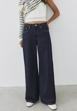 Person iført mørke, vidde jeans, hvide sko og en kort hvid top med et langærmet, stribet tøjdelemæssigt delvist draperet over den ene skulder.