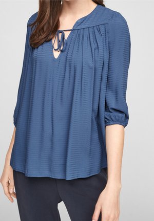 Vrouw draagt een losse blauwe blouse met textuur, een strik bij de halslijn en driekwart mouwen, gecombineerd met een donkere broek.