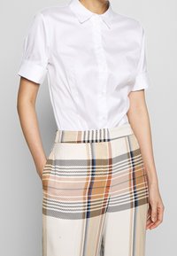 Wit overhemd met kraag en korte opgerolde mouwen, gecombineerd met beige geruite broek met hoge taille met zwarte, bruine en blauwe accenten.