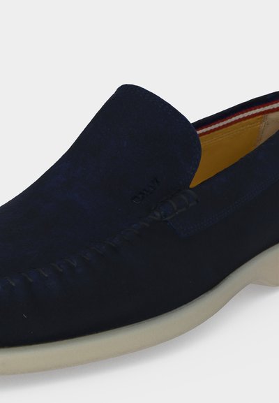 Μοκασίνι slip-on από σουέτ σε ναυτικό μπλε με λευκή σόλα, ορατά ραφές και κίτρινη εσωτερική επένδυση με κόκκινη, λευκή και μπλε διακόσμηση.