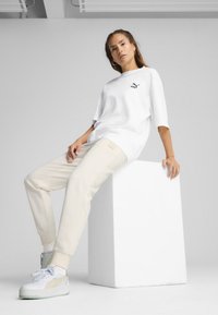 T-shirt bianca oversize con logo nero, pantaloni jogger crema con caviglie elastiche e sneaker bianche con dettagli verdi. Design minimalista.