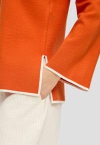 Oranje wolmix top met witte details, voorzien van een split hem en uitlopende mouwen. Hand geplaatst in een pocket op witte geribde broek.