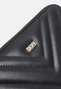 Coin de portefeuille en cuir noir avec couture en chevron et logo doré DKNY près d'une fermeture éclair dorée.