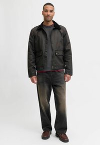 Brązowa woskowana kurtka z czarnym polarem przy kołnierzu, z dwoma przednimi kieszeniami, noszona na szarym podkoszulku i wyblakłych czarnych dżinsach, w połączeniu z brązowymi butami.