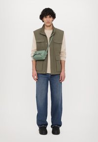 Gilet verde senza maniche sopra un maglione beige, abbinato a jeans a gamba larga blu e scarpe nere. È indossata una piccola borsa a tracolla verde.