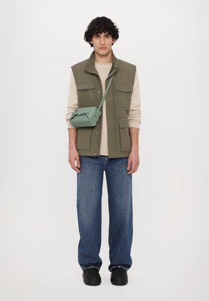 Vesta verde fără mâneci peste un pulover bej, asociată cu blugi largi albastri și pantofi negri. Se poartă o geantă mică verde tip crossbody.