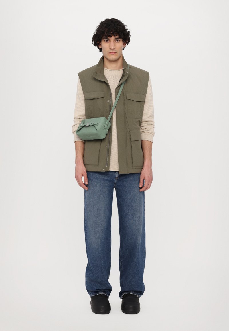 Gilet verde senza maniche sopra un maglione beige, abbinato a jeans a gamba larga blu e scarpe nere. È indossata una piccola borsa a tracolla verde.