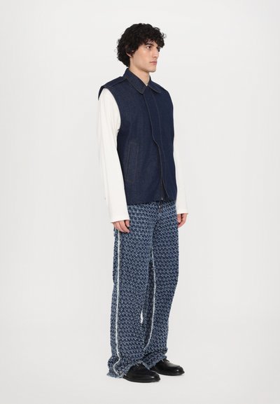 Denim vest with a collar and front closure, paired with textured, patterned, wide-leg trousers in blue and white, worn with black shoes.
