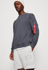 Szary sweatshirt z okrągłym dekoltem, z zamykaną na zamek kieszenią na rękawie i czerwoną metką. W zestawie z białymi szortami z tłoczonymi akcentami.