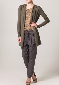 Olivgrön kofta, beige mönstrad topp, grå byxor med dragsko i midjan och volangdetaljer, matchat med bruna klackskor.