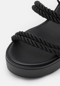 Sandale à plateforme noire avec des brides en corde torsadée, un dessus en simili cuir lisse et une semelle épaisse et arrondie.