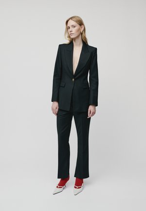 Black tailored suit with a deep V-neck blazer, slim pants, and pointed white heels paired with red socks. Gold-button detail on the blazer.
