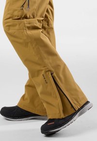 Beanie-Hosen aus leicht braunem Stoff mit seitlichen Reißverschlüssen, kombiniert mit schwarzen strukturierten Schuhen mit grauer Sohle und orangen Akzenten.