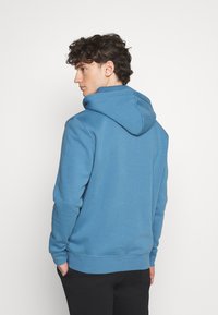 Felpa blu realizzata in tessuto morbido, dotata di un ampio cappuccio, polsini a costine e una vestibilità rilassata. Indossata con le mani nelle tasche e pantaloni neri.