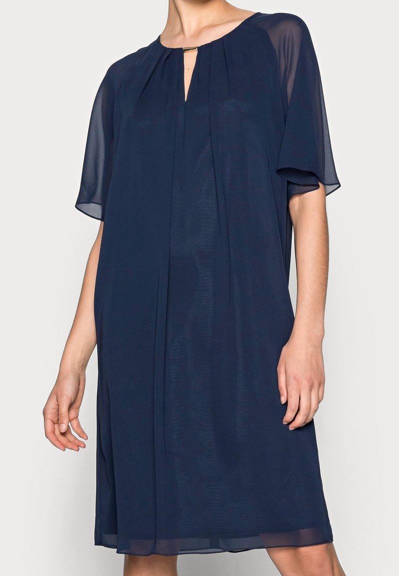 Robe bleu marine en tissu transparent, avec manches courtes, encolure ronde avec un détail en trou de serrure, et des plis doux pour une coupe fluide.