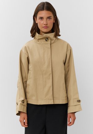 Femme aux cheveux bruns mi-longs, portant une veste beige boutonnée à col montant et un pantalon noir, sur un fond uni.