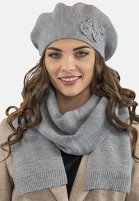 Un boina de punto gris presenta un acento floral y una bufanda gris a juego con bordes acanalados, usada sobre un abrigo beige. Texturas suaves y cómodas.