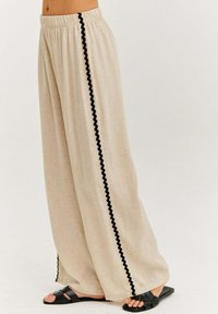 Béžové kalhoty širokého střihu s texturovanou tkaninou, s černým ozdobným páskem po straně a elastickým pasem. Pod nimi ploché černé sandály.