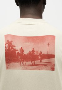 Τρεις καουμπόηδες με καπέλα καβαλούν άλογα σε ένα χωματόδρομο με δέντρα και κολόνα ηλεκτρικού ρεύματος στο φόντο, τυπωμένοι σε μια μπεζ μπλούζα.