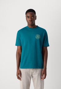 Teal bomuld t-shirt med rund halsudskæring, korte ærmer og et trykt logo på brystet med et tennismotiv. Simpelt design, blød tekstur.