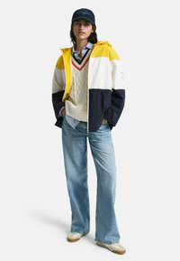 Giacca bicolore gialla, bianca e blu navy, sovrapposta a un maglione lavorato a maglia crema e una camicia a righe blu, abbinata a jeans denim chiari.