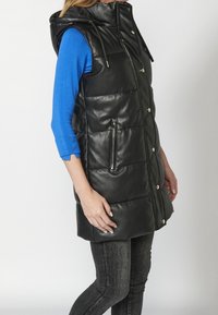 Femme portant un gilet sans manches noir matelassé à capuche par-dessus un haut bleu à manches longues et un jean gris foncé, debout de profil.