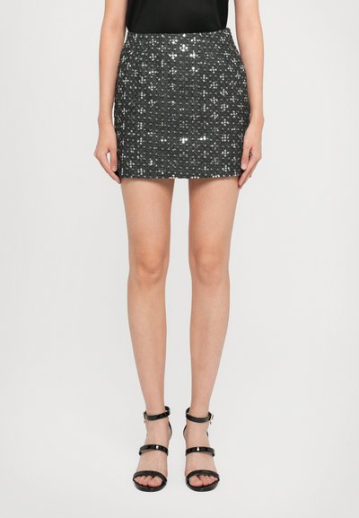 Donna che indossa una mini gonna nera di paillettes, un top nero senza maniche e sandali alti con cinturini neri, in piedi contro uno sfondo bianco.