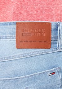 Ljusblå denim med en brun lädertagg som står "HILFIGER DENIM" och "AN AMERICAN ORIGINAL." Har sömmar och en liten logotypaccent.