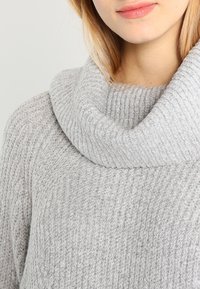 Grå stickad tröja med en tjock, vikbar krage. Har en vertikal ribbstruktur. Mjuk tyg med ett lätt, något fläckigt utseende.