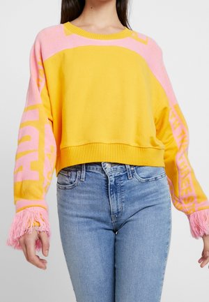 Kobieta w krótkim żółtym swetrze z różowym napisem na rękawach i różowymi frędzlami na mankietach, w połączeniu z wysokim stanem niebieskich dżinsów z ćwiekami po bokach.