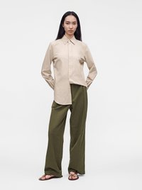 Femme debout portant une chemise beige à manches longues boutonnée, un pantalon large olive et des sandales plates marron sur un fond blanc uni.