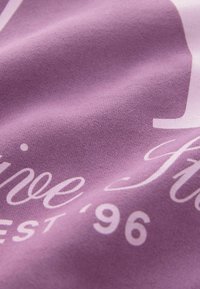 Gros plan sur un tissu violet avec un texte blanc imprimé en cursive et en bloc montrant partiellement « Fine » et « est '96 ».