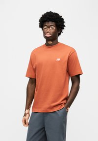 Πορτοκαλί βαμβακερό t-shirt με κοντά μανίκια και στρογγυλή λαιμόκοψη, με μικρό λευκό λογότυπο στο αριστερό στήθος, συνδυασμένο με γκρι σορτς.