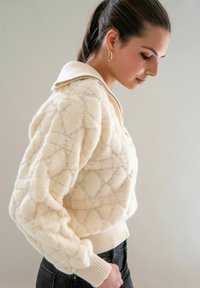 Crème trui met een gestructureerd ruitpatroon, een ritskraag en geribde boorden. Gedragen over zwarte jeans, met een gladde stof en een getailleerde pasvorm.