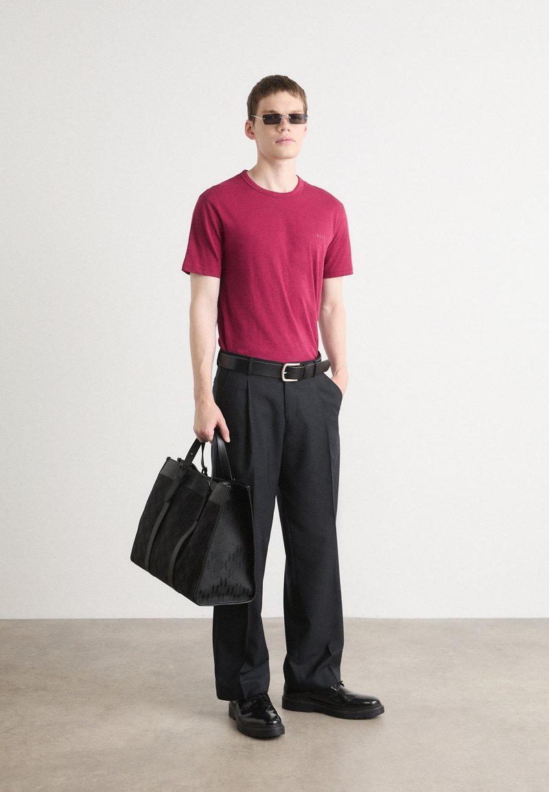 T-shirt en coton bordeaux, pantalon noir ajusté, chaussures en cuir noires, sac fourre-tout noir à motifs et lunettes de soleil sombres. Fond uni.