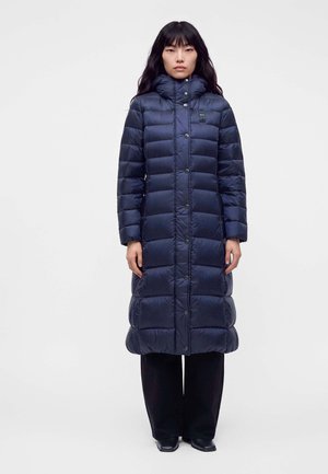 Femme aux longs cheveux noirs portant un long manteau matelassé bleu marine à col montant, un pantalon noir et des chaussures noires, debout devant un fond blanc.